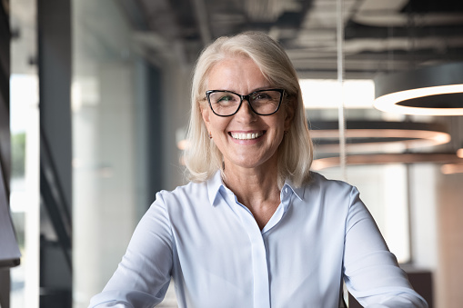 Headshot of smiling middle-aged businesswoman in glasses look at camera pose shooting live webinar broadcast in office, happy mature female employee talk on video call at workplace, leadership concept